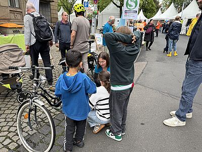 Im Vordergrund stehen 2 Kinder vor einem Fahrrad, dahinter kniet Patricia mit einem dritten Kind. Im Hintergrund Erwachsene , Teils mit Fahrradhelm und der Stand des VCD