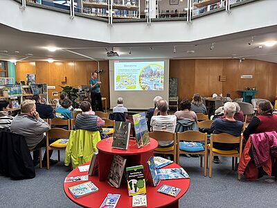 In einer kreisförmigen Bibliothek sitzen mehrere Personen im Publikum und verfolgen einen Vortrag eines Mannes vor einer Präsentationsleinwand zum Thema „Barcelona“. Im Vordergrund steht ein roter runder Buchtisch, auf dem verschiedene Bücher zum Thema Nachhaltigkeit und Mobilität ausgestellt sind.