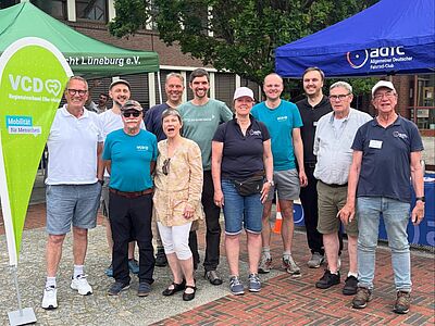Eine Gruppe von elf Personen steht lächelnd im Freien auf einem Pflaster- und Backsteinboden. Sie tragen Freizeitkleidung (T-Shirts und kurze Hosen). Im Hintergrund sind ein grünes Zelt („VCD Regionalverband Elbe-Heide“), ein blaues Zelt („ADFC“) sowie ein Wohngebäude und Bäume zu sehen. Links im Vordergrund steht eine große, grüne, tropfenförmige VCD-Fahne.