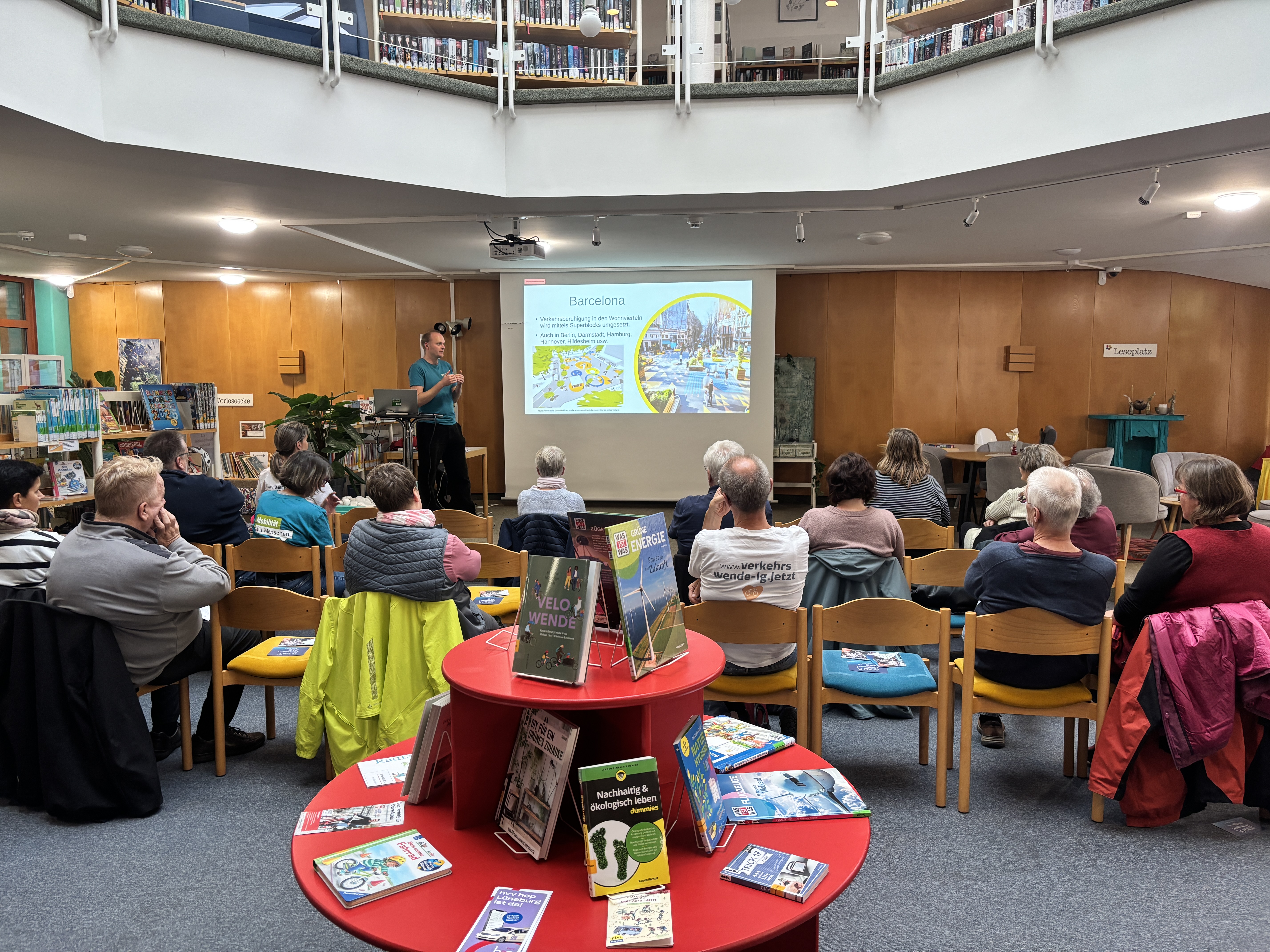 In einer kreisförmigen Bibliothek sitzen mehrere Personen im Publikum und verfolgen einen Vortrag eines Mannes vor einer Präsentationsleinwand zum Thema „Barcelona“. Im Vordergrund steht ein roter runder Buchtisch, auf dem verschiedene Bücher zum Thema Nachhaltigkeit und Mobilität ausgestellt sind.