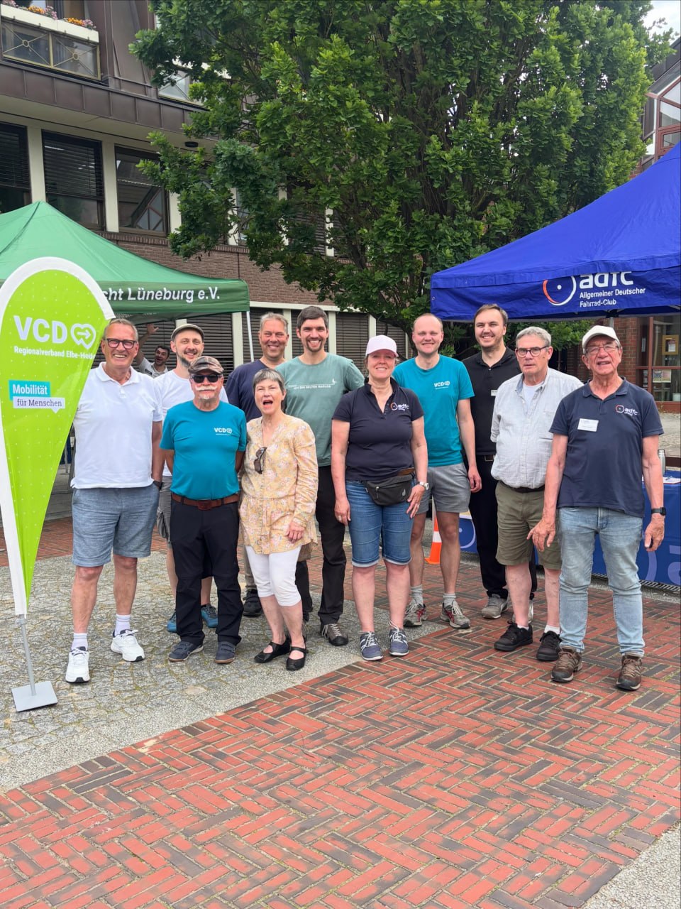 Eine Gruppe von elf Personen steht lächelnd im Freien auf einem Pflaster- und Backsteinboden. Sie tragen Freizeitkleidung (T-Shirts und kurze Hosen). Im Hintergrund sind ein grünes Zelt („VCD Regionalverband Elbe-Heide“), ein blaues Zelt („ADFC“) sowie ein Wohngebäude und Bäume zu sehen. Links im Vordergrund steht eine große, grüne, tropfenförmige VCD-Fahne.